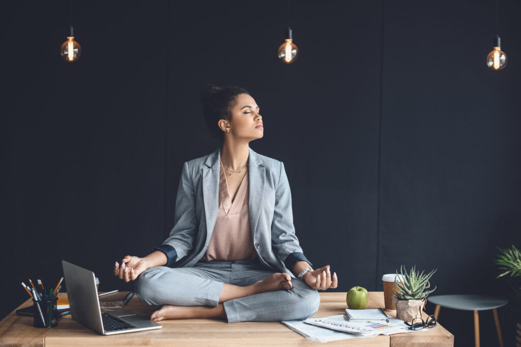 African American businesswoman sitting in lotus pose on the desk while meditating in office.
